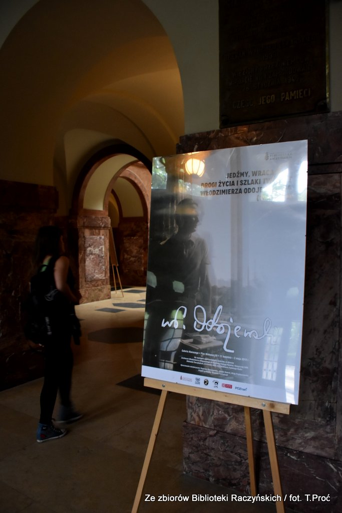 W holu Collegium Maius stoi sztaluga z plakatem promującym wystawę poświęconą Odojewskiemu, obok przechodząca studentka.