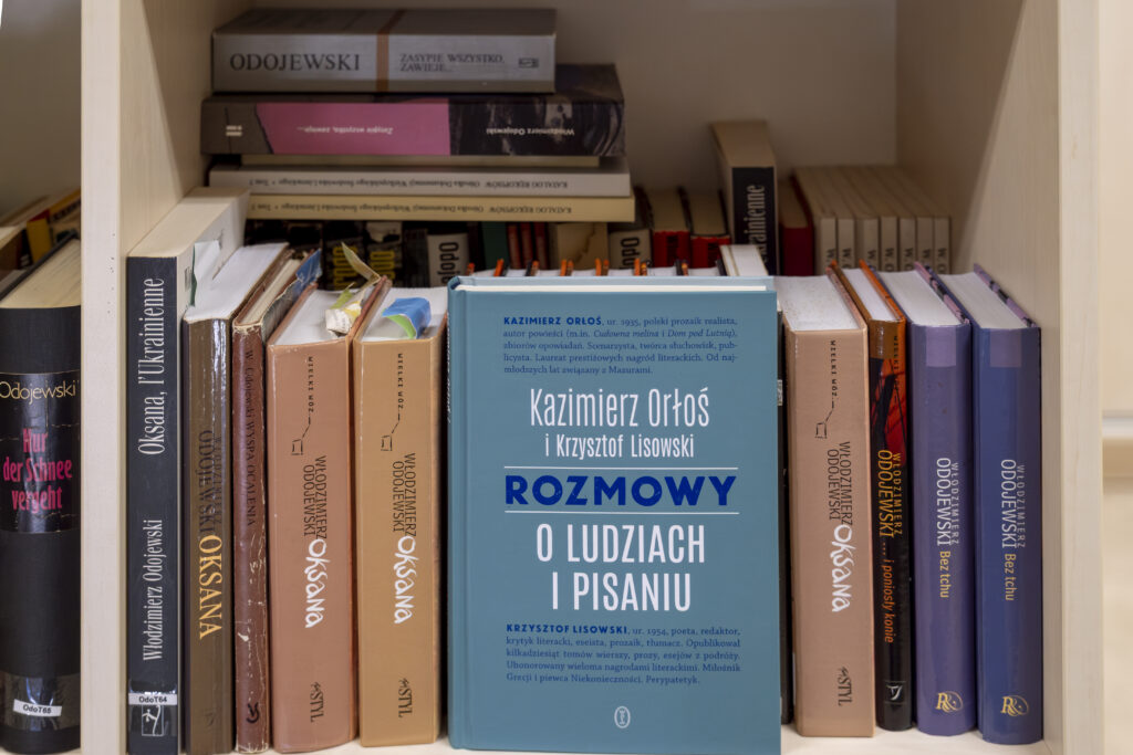 Na półce drewnianego regału stoją w dwóch rzędach wydania powieści Włodzimierza Odojewskiego, pośród nich przodem okładki skierowana do obiektywu książka Kazimierza Orłosia i Krzysztofa Lisowskiego pt. „Rozmowy o pisarzach i pisaniu” Wydawnictwa Literackiego.