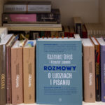 Na półce drewnianego regału stoją w dwóch rzędach wydania powieści Włodzimierza Odojewskiego, pośród nich przodem okładki skierowana do obiektywu książka Kazimierza Orłosia i Krzysztofa Lisowskiego pt. „Rozmowy o pisarzach i pisaniu” Wydawnictwa Literackiego.